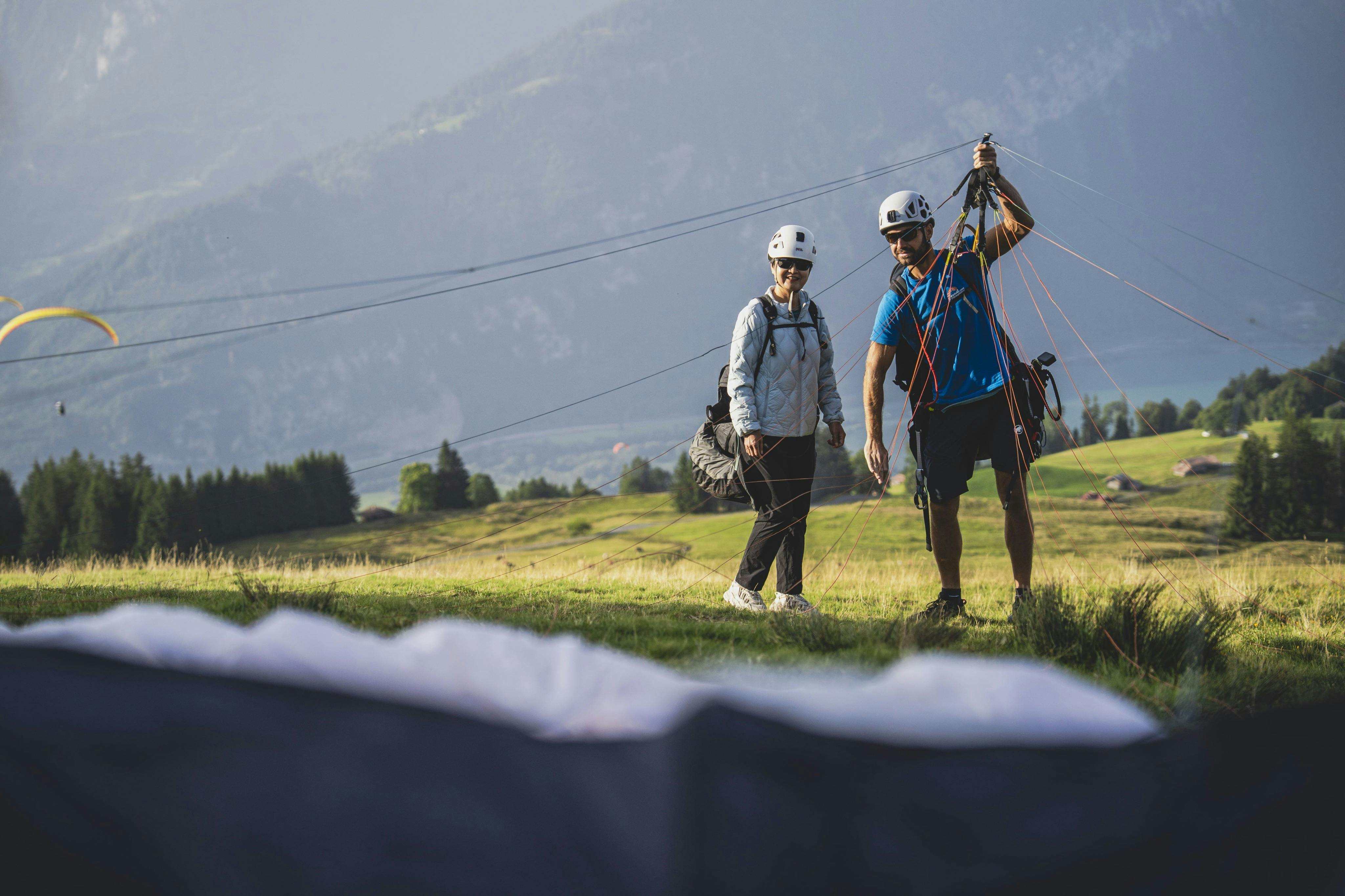 Deux personnes portant des casques se tiennent dans un champ herbeux avec un équipement de parapente dans une zone montagneuse.