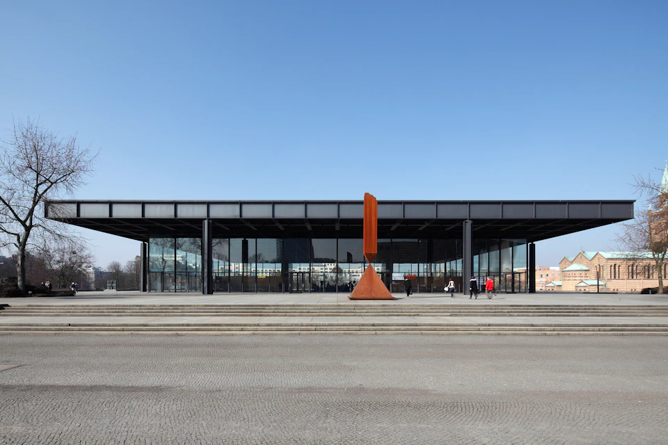 Modern glass building with a large courtyard, featuring a tall, rust-colored triangular sculpture, under a clear blue sky.