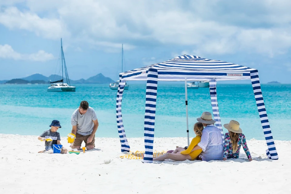 Family under a striped canopy on a white sand beach; father and child building sandcastles; turquoise water and sailboats in background.