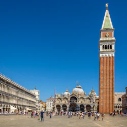 Ett livligt torg med ett högt klocktorn, utsmyckade kupolformade basilika och långa kolonnadade byggnader under en klarblå himmel.