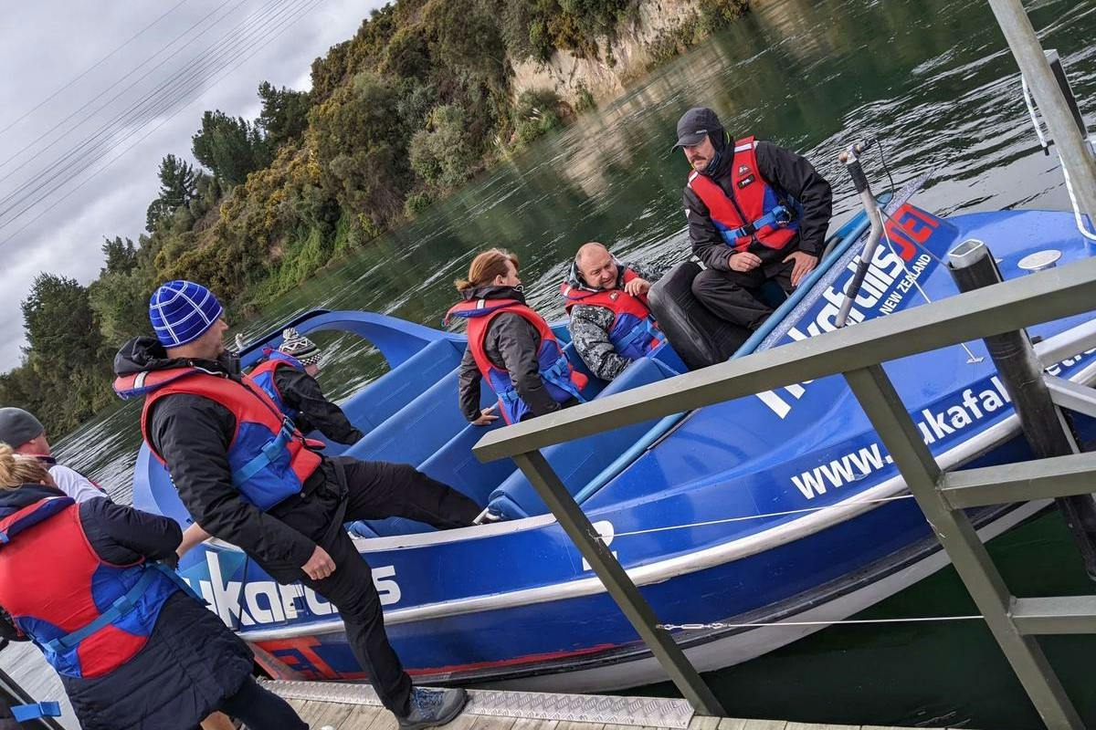 Four people wearing life jackets sit in a blue motorboat docked near a wooden platform on a calm waterway.
