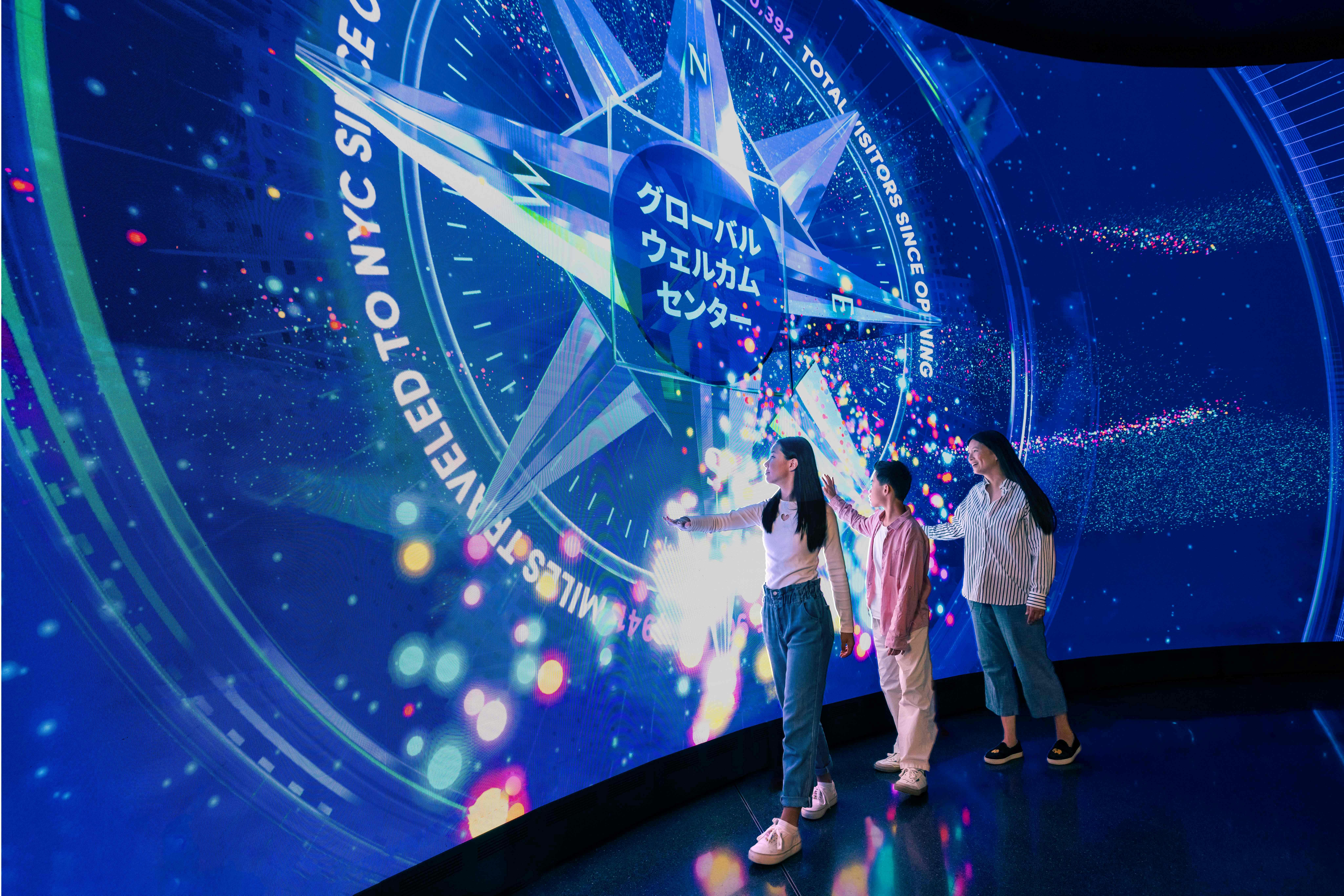 Three people interact with a large illuminated touchscreen displaying colorful graphics and Japanese text in a dark room.