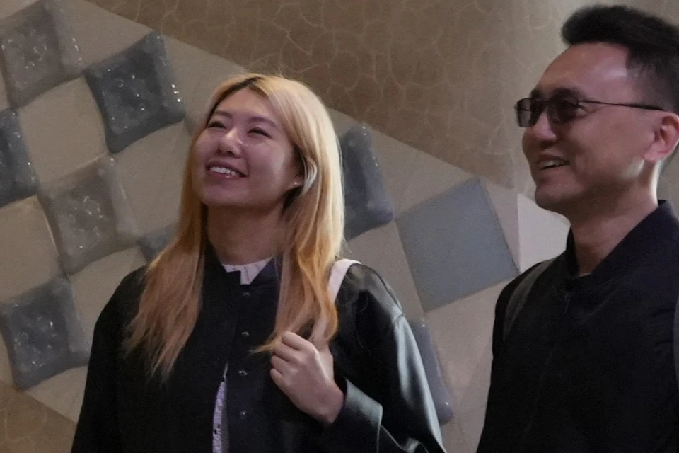 A group of people stand indoors smiling, with a patterned wall and staircase behind them. They carry white tote bags.