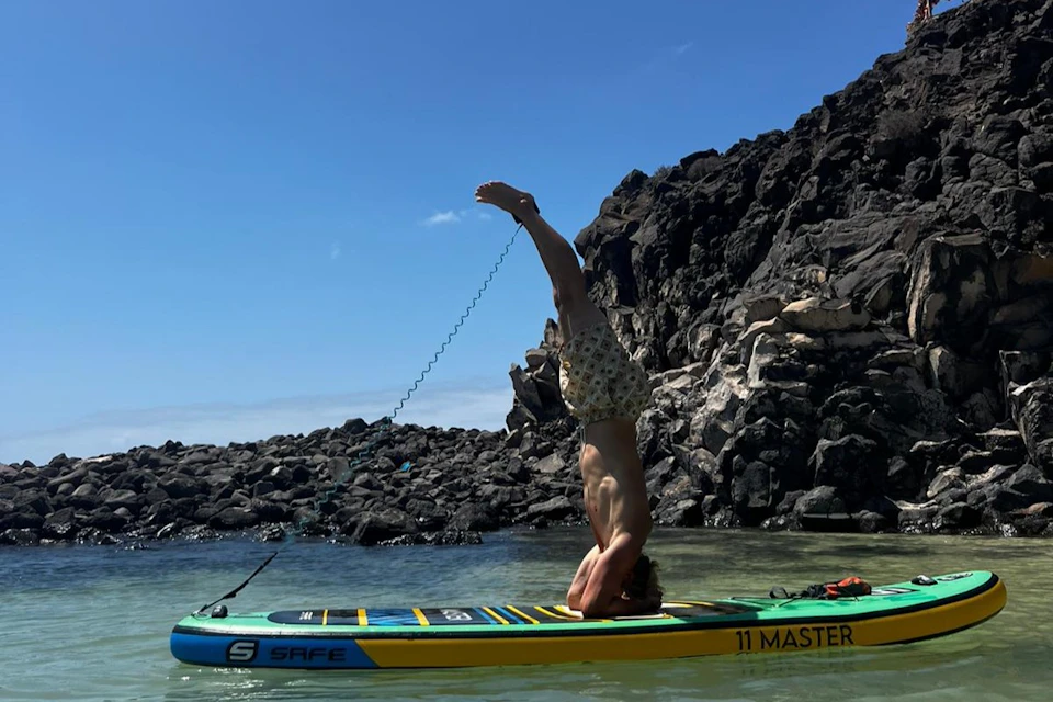 Person performing a headstand on a paddleboard in calm water near rocky shore under a clear blue sky.