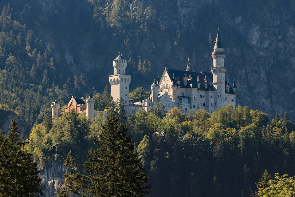 A castle with multiple towers sits atop a forested hill, surrounded by trees and backed by a mountain.