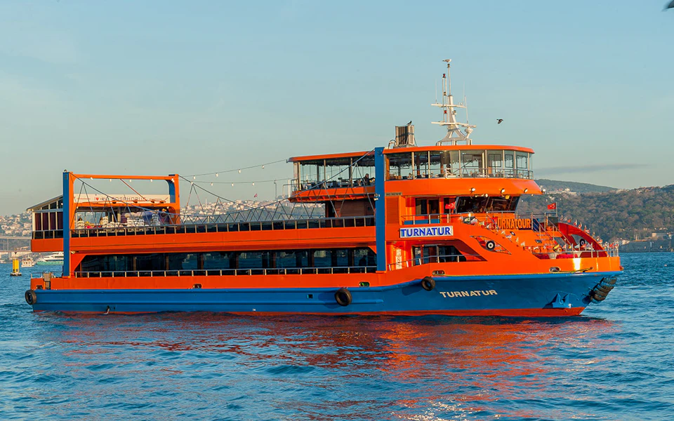 An orange and blue passenger ferry named "TURNATUR" on calm water with a cityscape in the distant background.