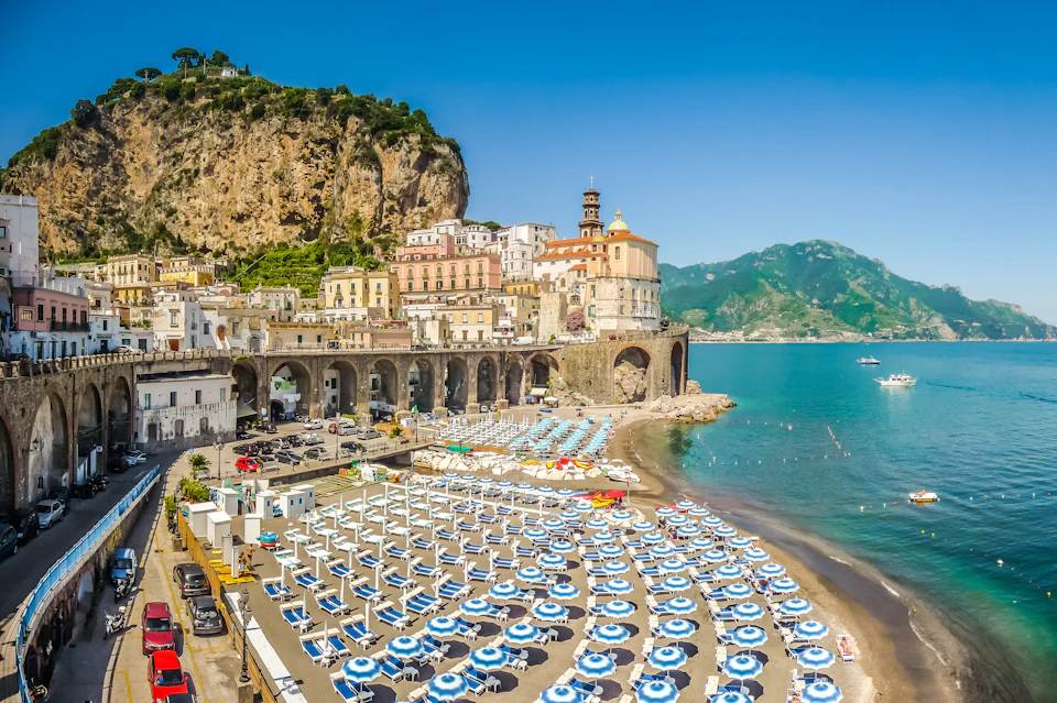 A coastal town with colorful buildings on a cliff, a beach filled with blue umbrellas, and mountains in the background.