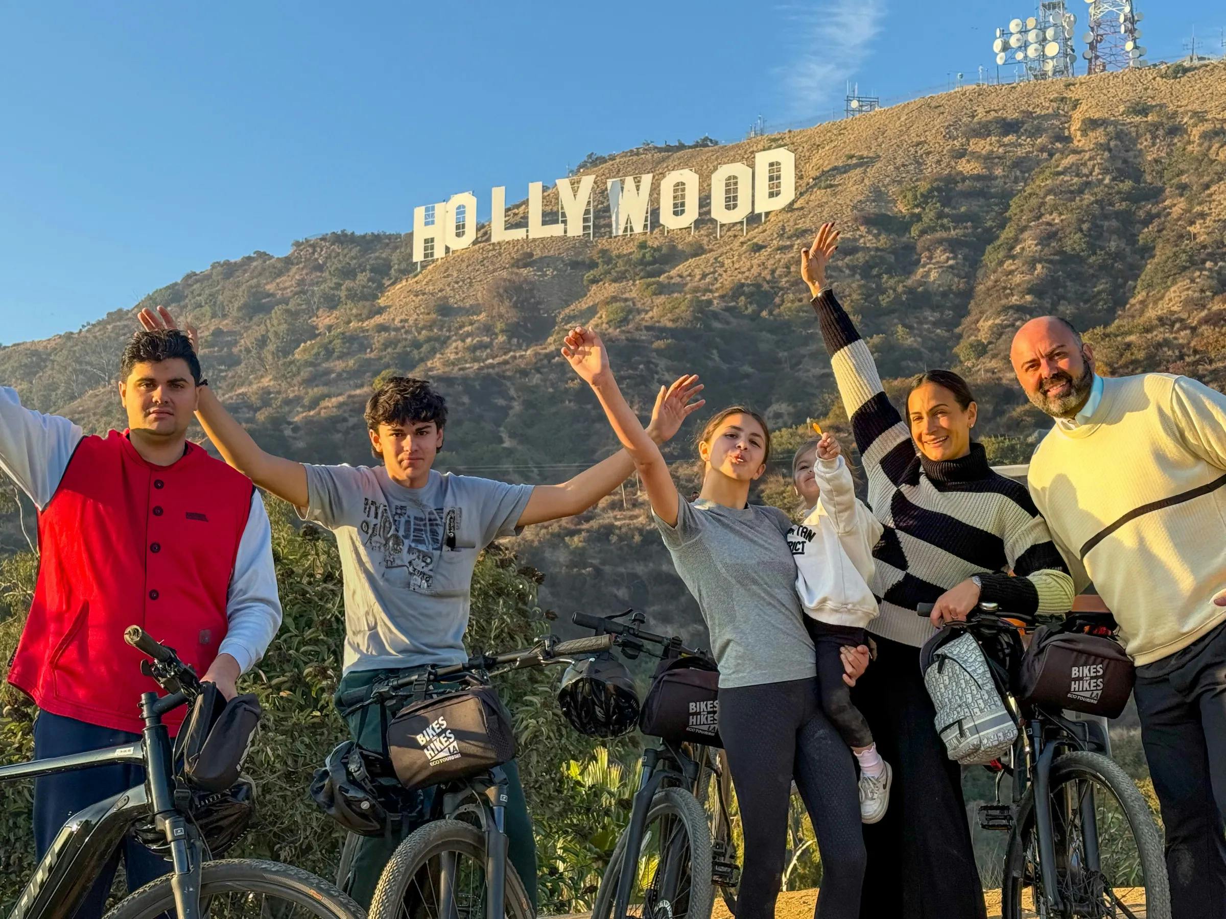 Five people posing with bicycles in front of the Hollywood sign on a sunny day, raising their arms in celebration.