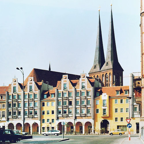 Serie av sammanhängande byggnader med varierande fasader och två höga kyrkspiror i bakgrunden. Bilar parkerade framför.