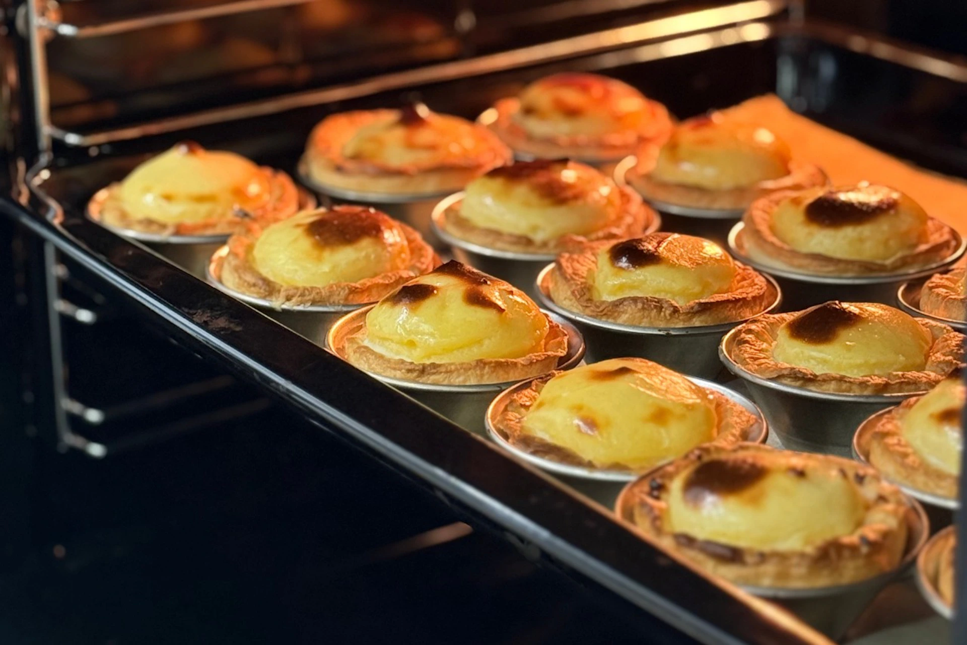 Rows of golden egg tarts baking in an oven, displayed on metal trays.
