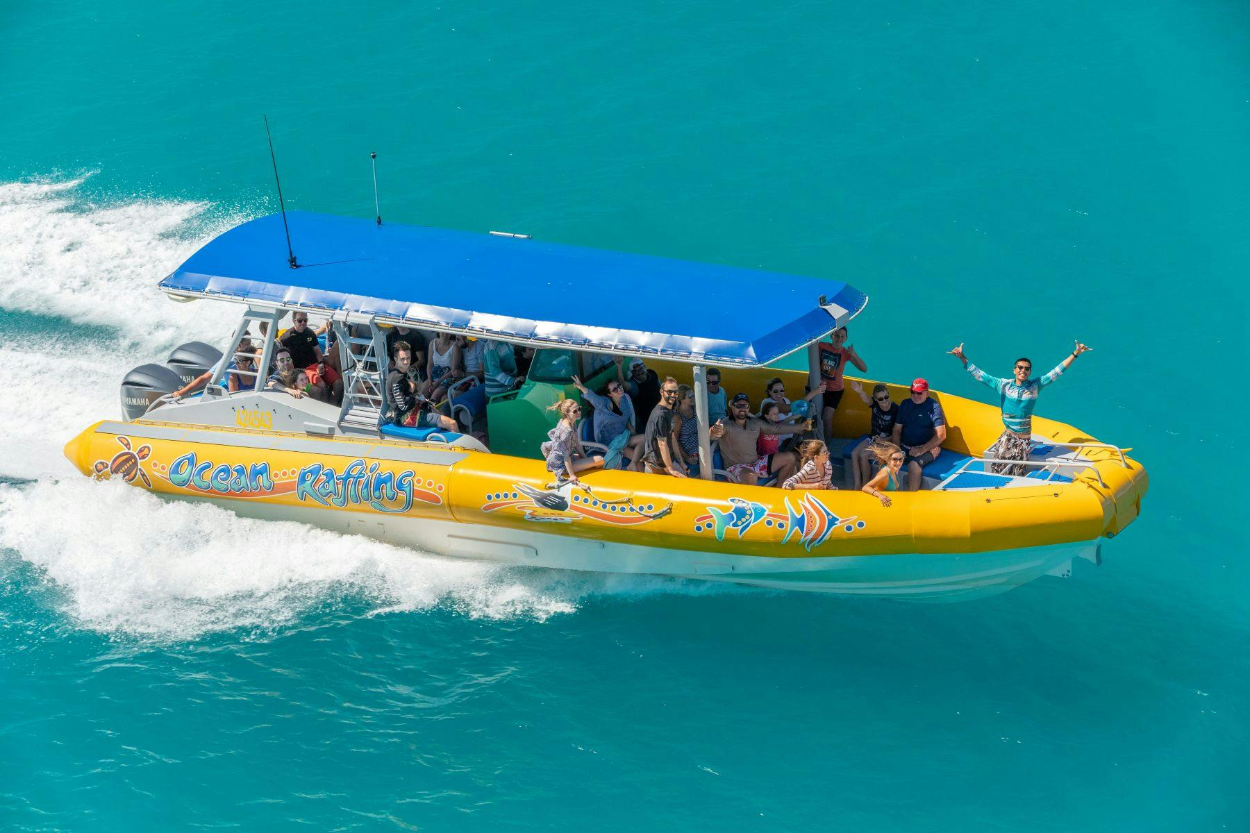 A yellow boat with a blue canopy on turquoise water, carrying passengers and a guide standing with arms raised.
