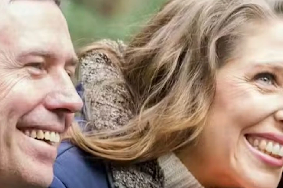 A man and a woman leaning on a railing outdoors, smiling, surrounded by trees and greenery.