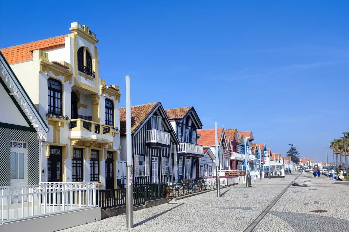 Πολύχρωμα σπίτια με ριγέ προσόψεις και μπαλκόνια πλαισιώνουν έναν πλακόστρωτο δρόμο κάτω από έναν καταγάλανο ουρανό.