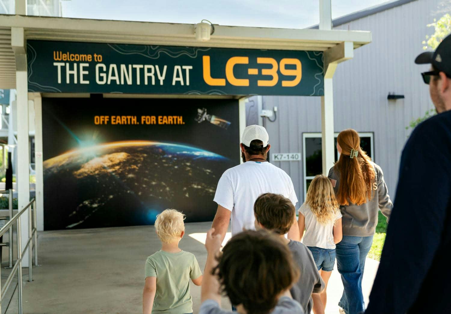 A family walks towards a building with a sign reading "THE GANTRY AT LC-39" and an image of Earth from space.