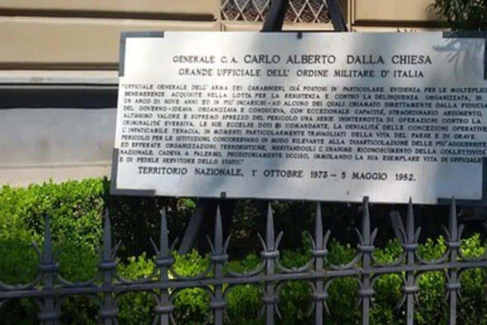 A metal plaque honoring Generale C.A. Carlo Alberto Dalla Chiesa, displayed behind a black iron fence with green foliage.