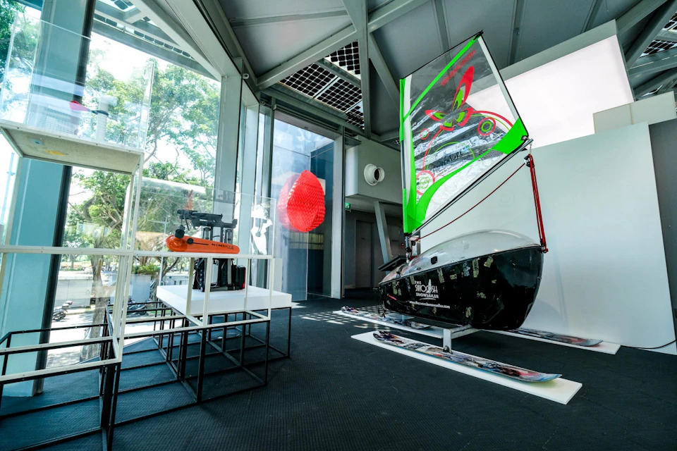 Indoor display featuring a sleek black boat with a vibrant green sail, a red sculpture, and various small mechanisms on tables.
