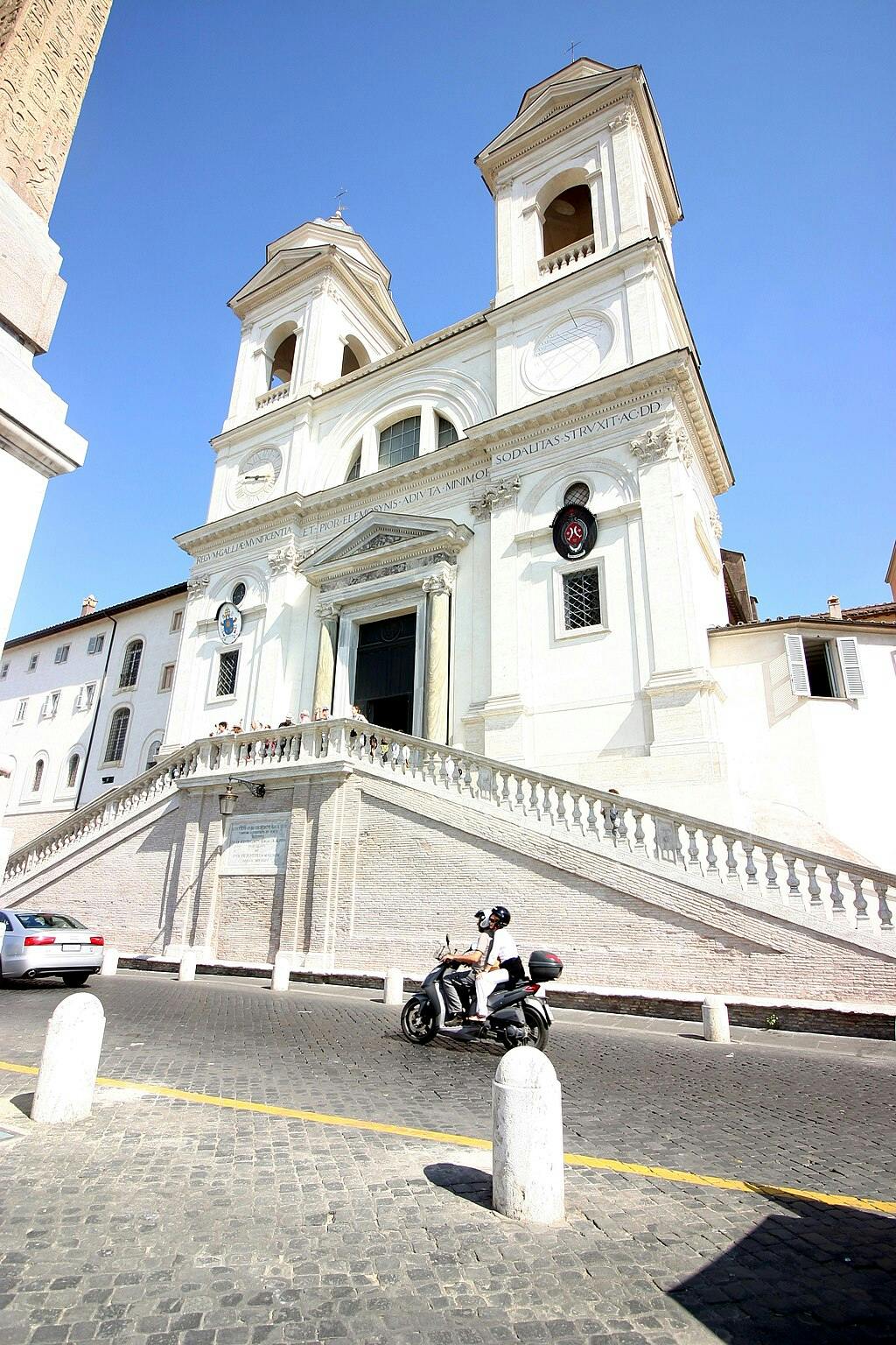 Igreja branca com torres sineiras gémeas, grande escadaria e pessoas nos degraus. Um homem anda de scooter numa rua de calçada.