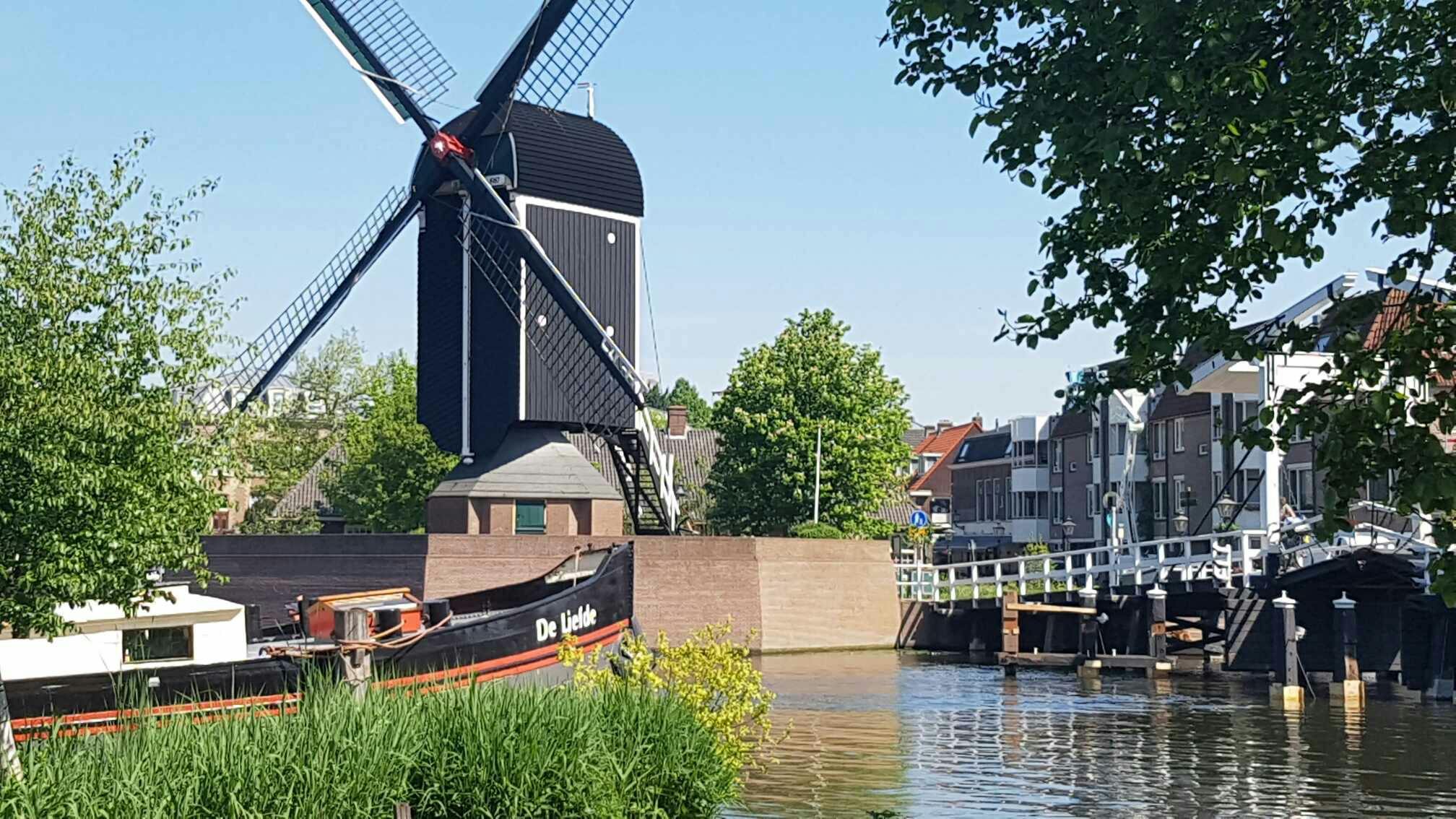 U kanálu stojí větrný mlýn s kotvícím člunem „De Liefde“, obklopený zelení a budovami pod jasnou oblohou.