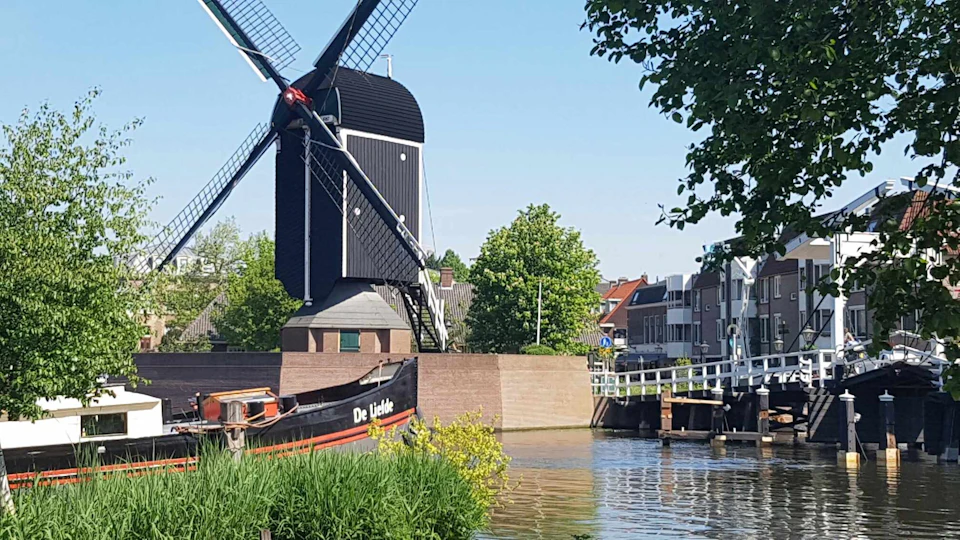 A windmill stands by a canal with a moored boat named "De Liefde," surrounded by greenery and buildings under a clear sky.