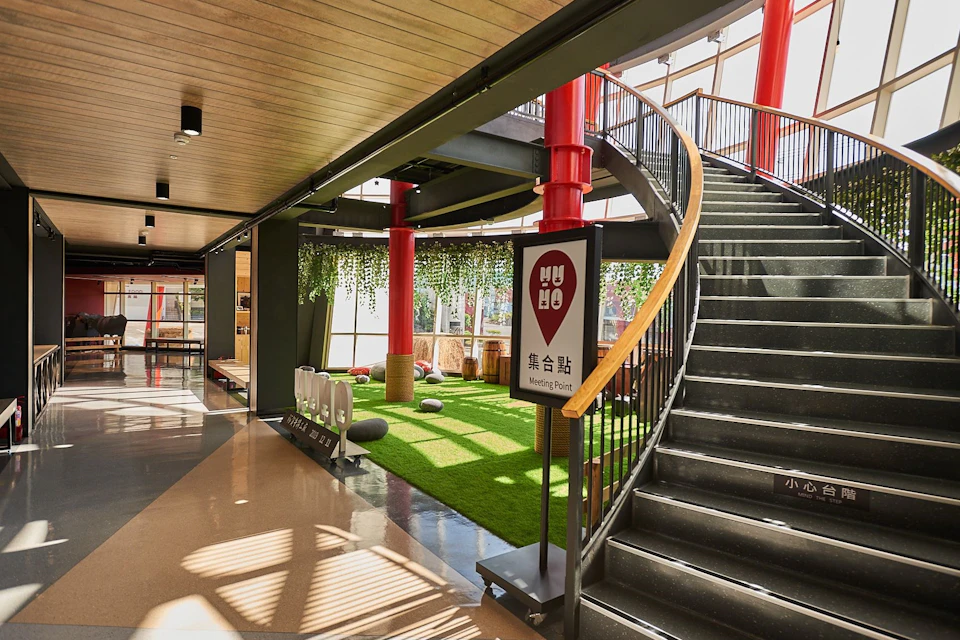 A modern indoor space with grass flooring, red pillars, a spiral staircase, and a sign reading 'Meeting Point'.