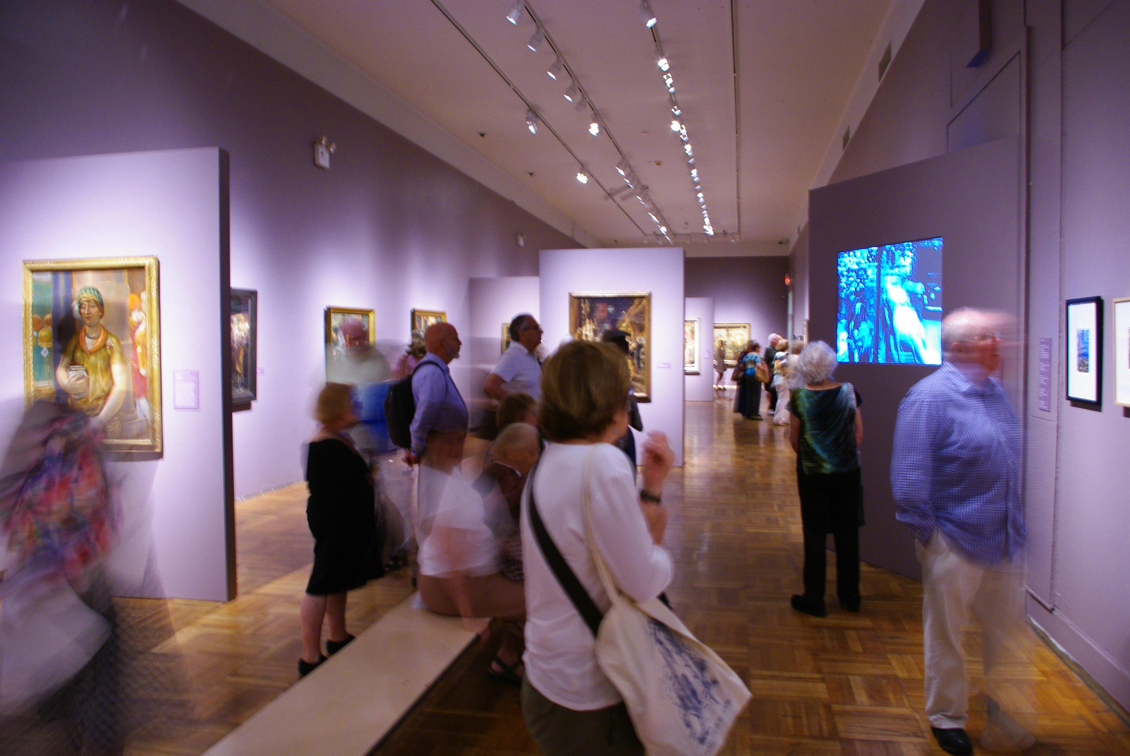 Άνθρωποι που θαυμάζουν διάφορα πλαισιωμένα έργα τέχνης σε μια έντονα φωτισμένη μουσειακή γκαλερί με τοίχους από λεβάντα και ξύλινα δάπεδα.