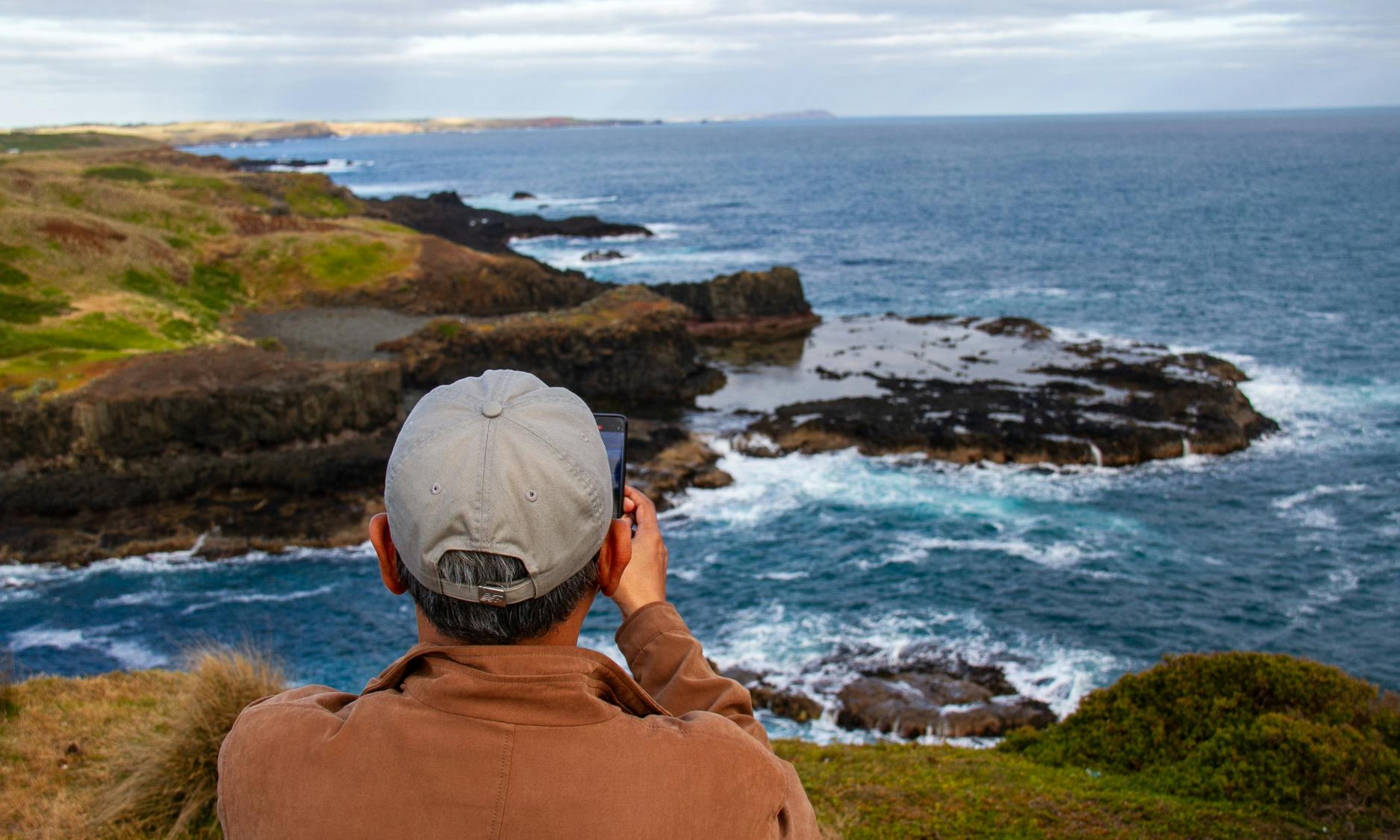 Osoba w szarej czapce robi zdjęcie skalistego wybrzeża i widoku na ocean.
