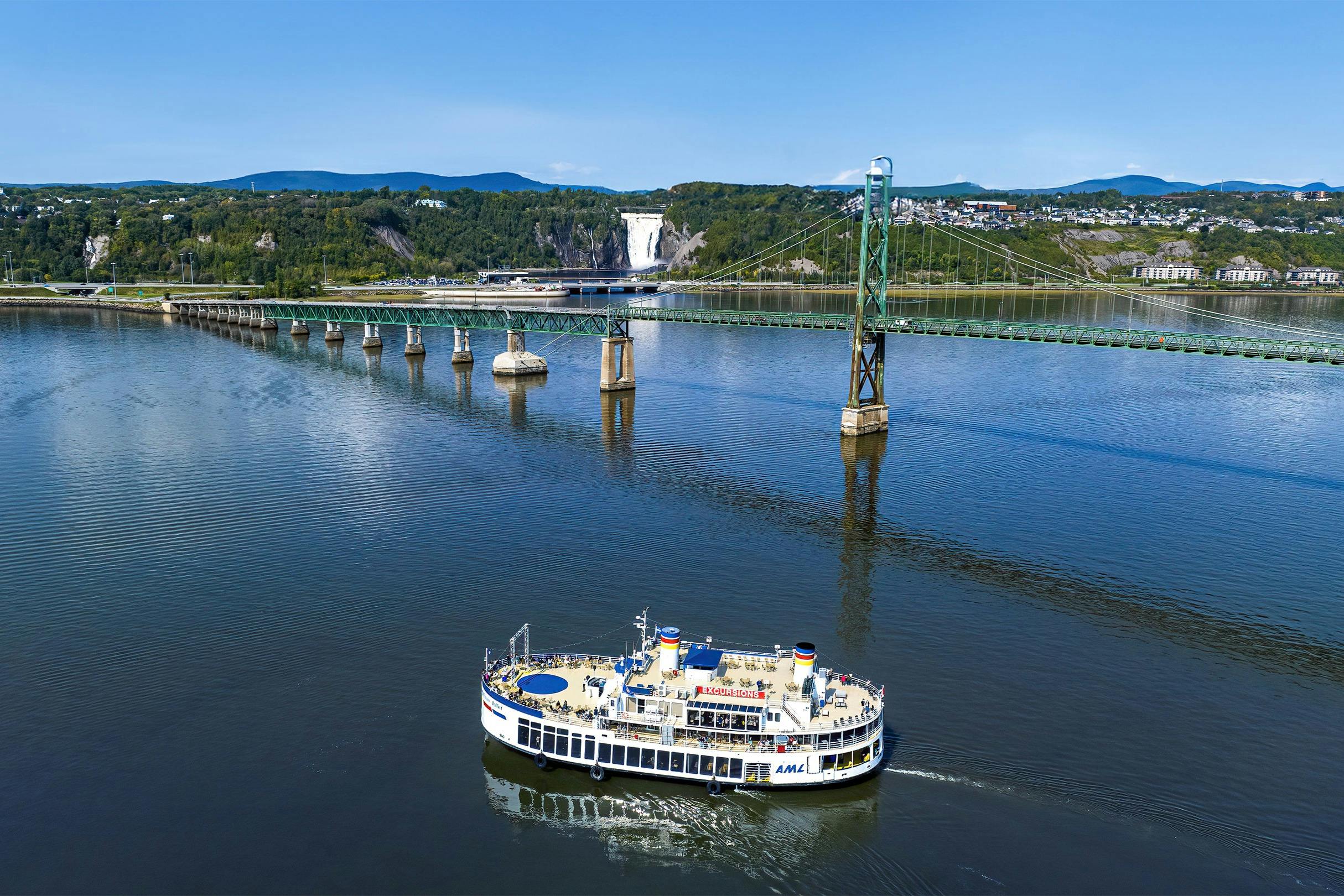 Ein Touristenboot fährt auf einem ruhigen Fluss in der Nähe einer Hängebrücke und eines Wasserfalls in einer üppigen, grünen Landschaft.
