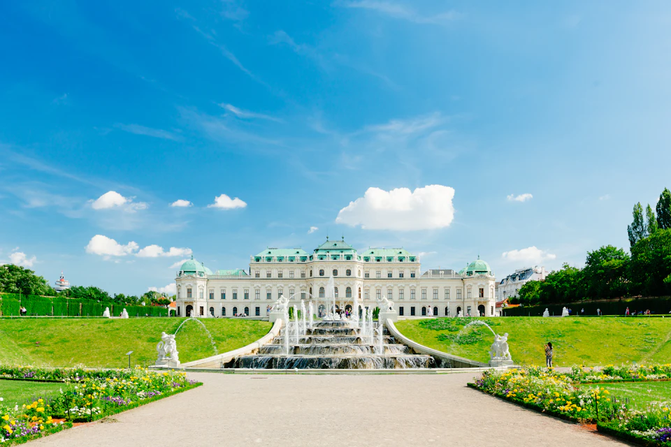 A grand white palace with green domes, surrounded by manicured gardens and fountains under a bright blue sky with scattered clouds.