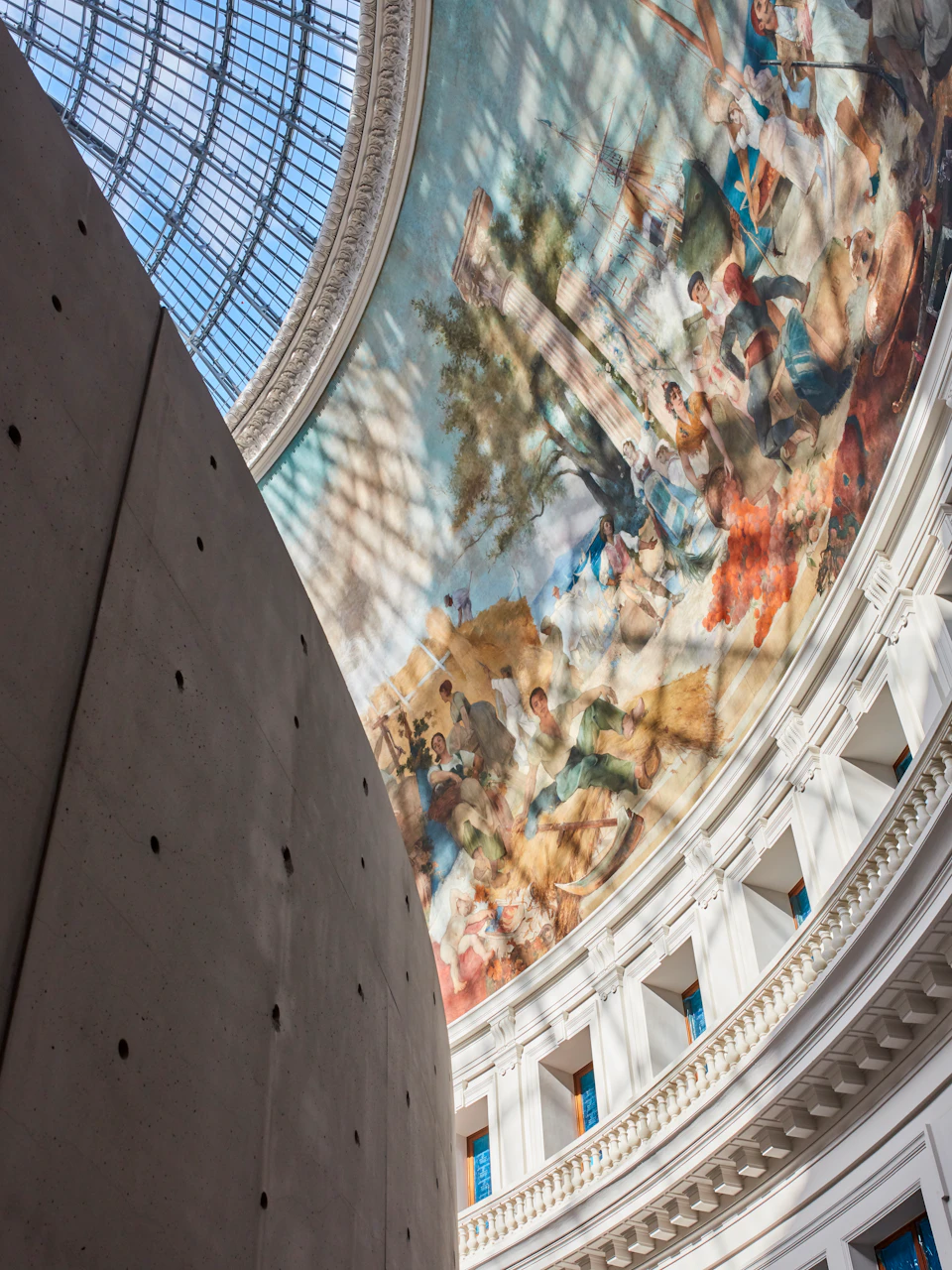 A curved wall inside a building with a large, detailed mural depicting people and nature under a domed, glass ceiling.