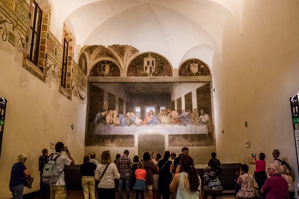A group of people viewing Leonardo da Vinci's "The Last Supper" mural in a large, arch-ceilinged room.
