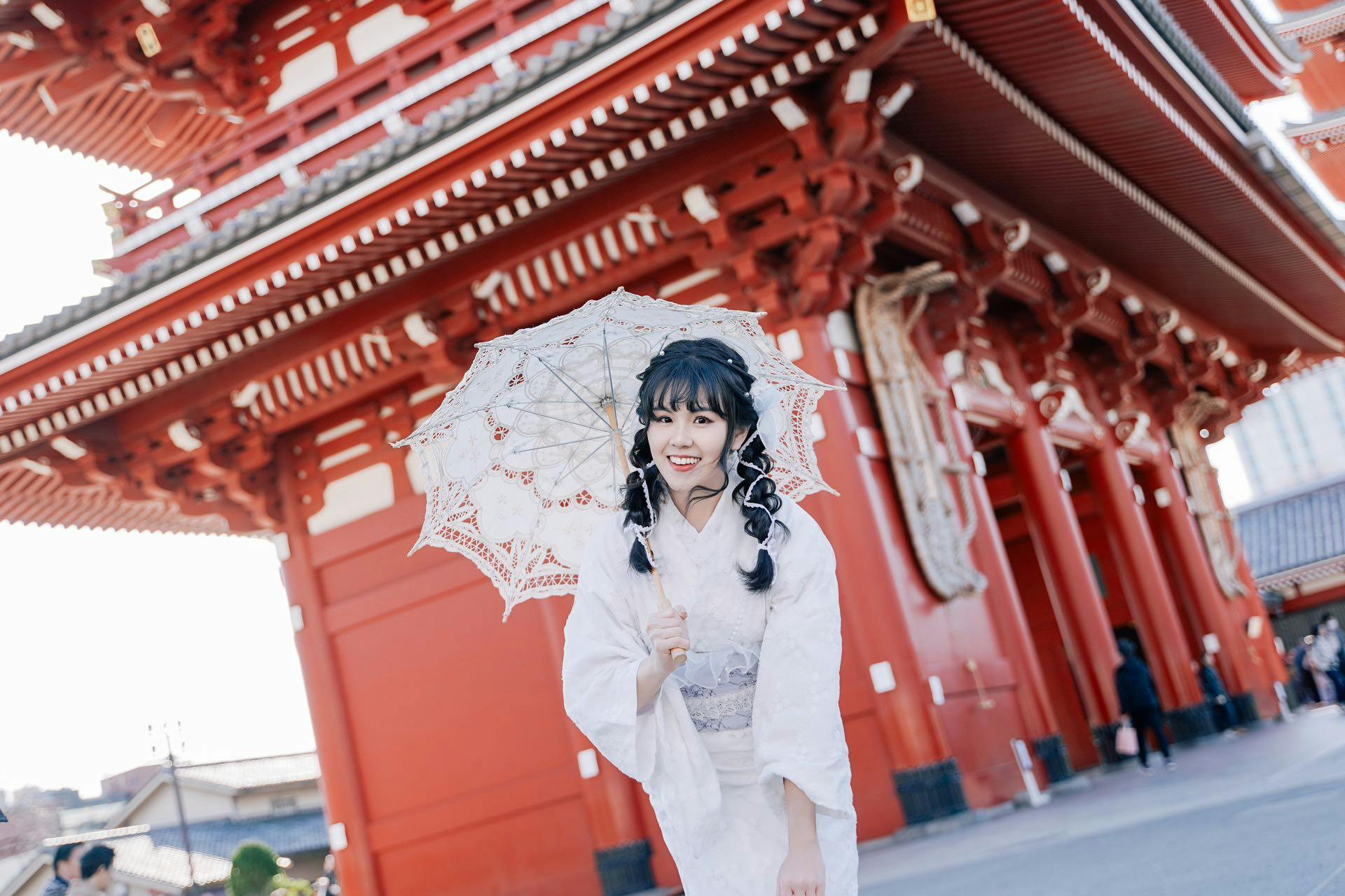 Person in traditional attire holding a lace parasol, standing in front of a red, ornate building.