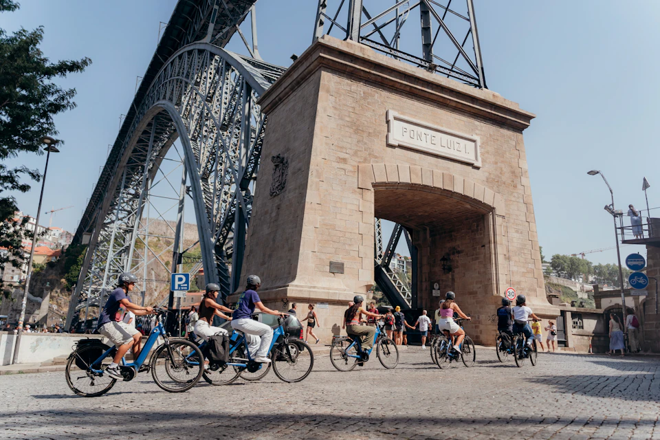 Cyclists and pedestrians passing under a stone arch of the Dom Luís I Bridge on a sunny day.