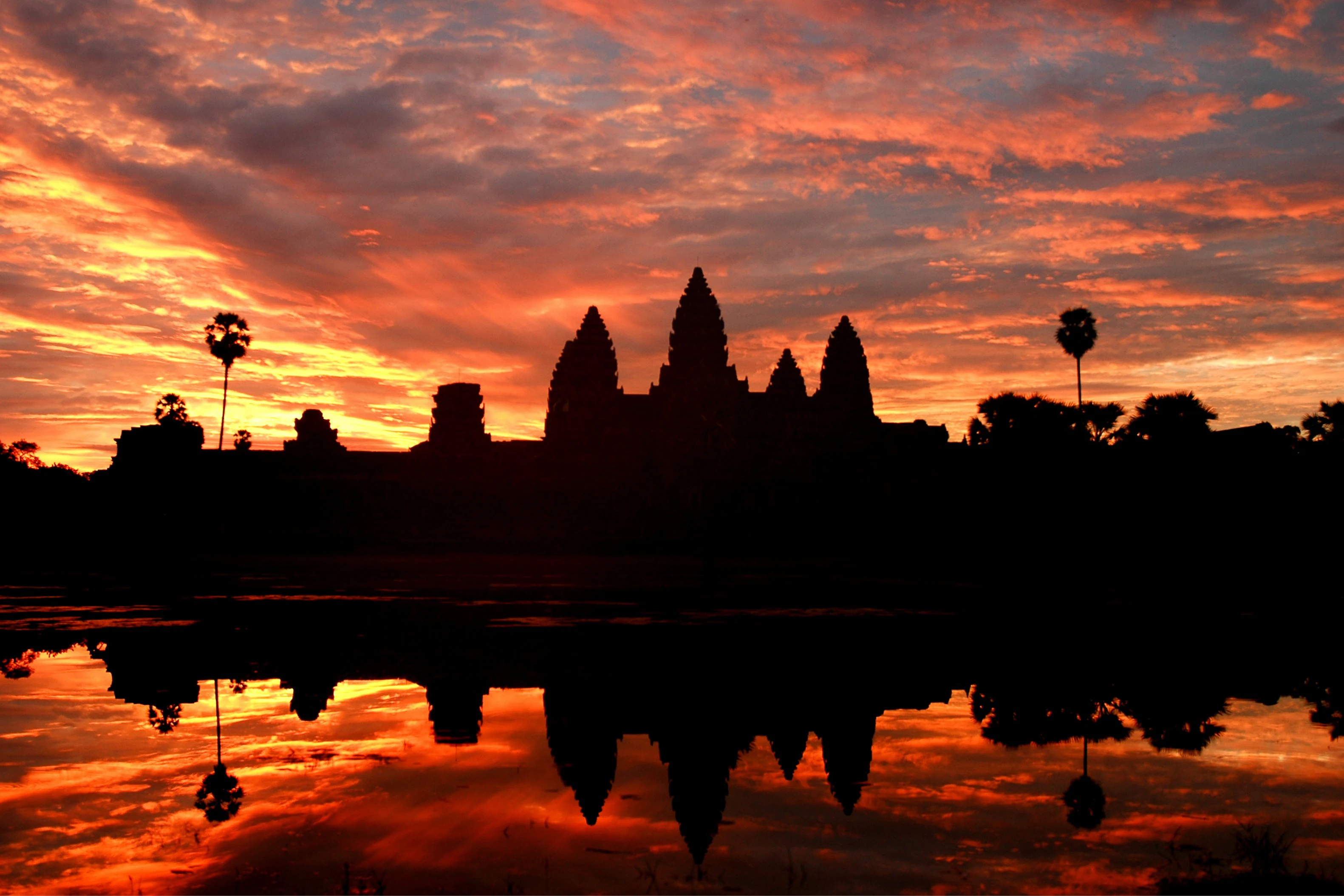 Silhouette of Angkor Wat against a vibrant orange and red sunrise, with its reflection in a calm body of water.