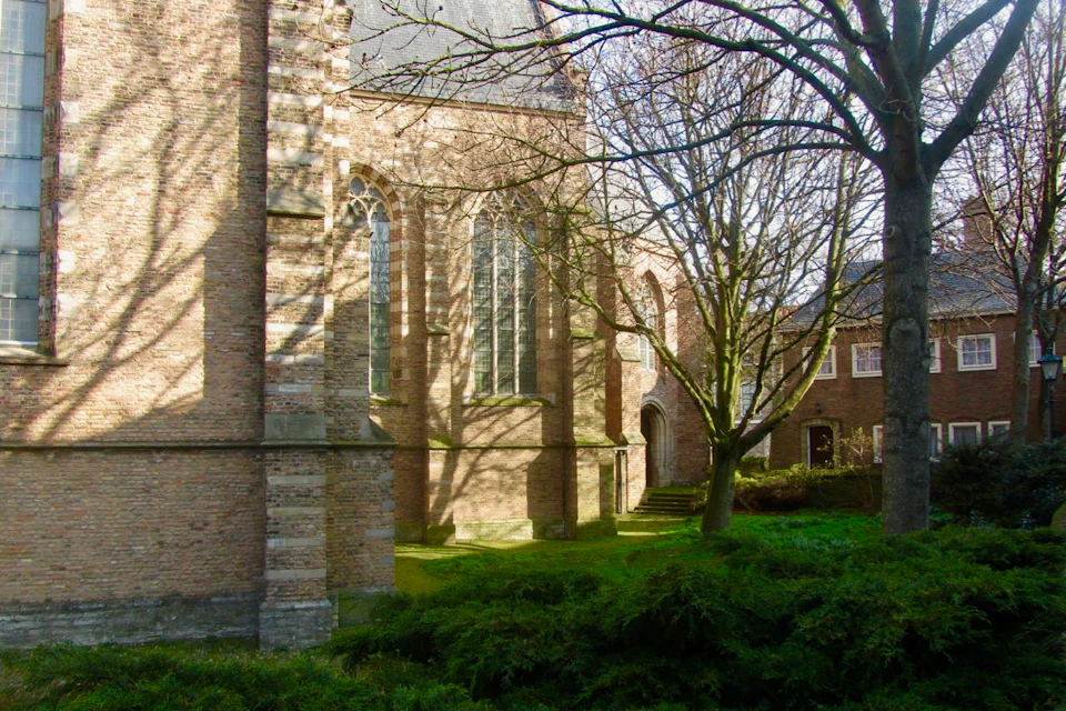 Sunlit brick church with arched windows, surrounded by bare trees and green bushes, casting shadows on the grass and structure.