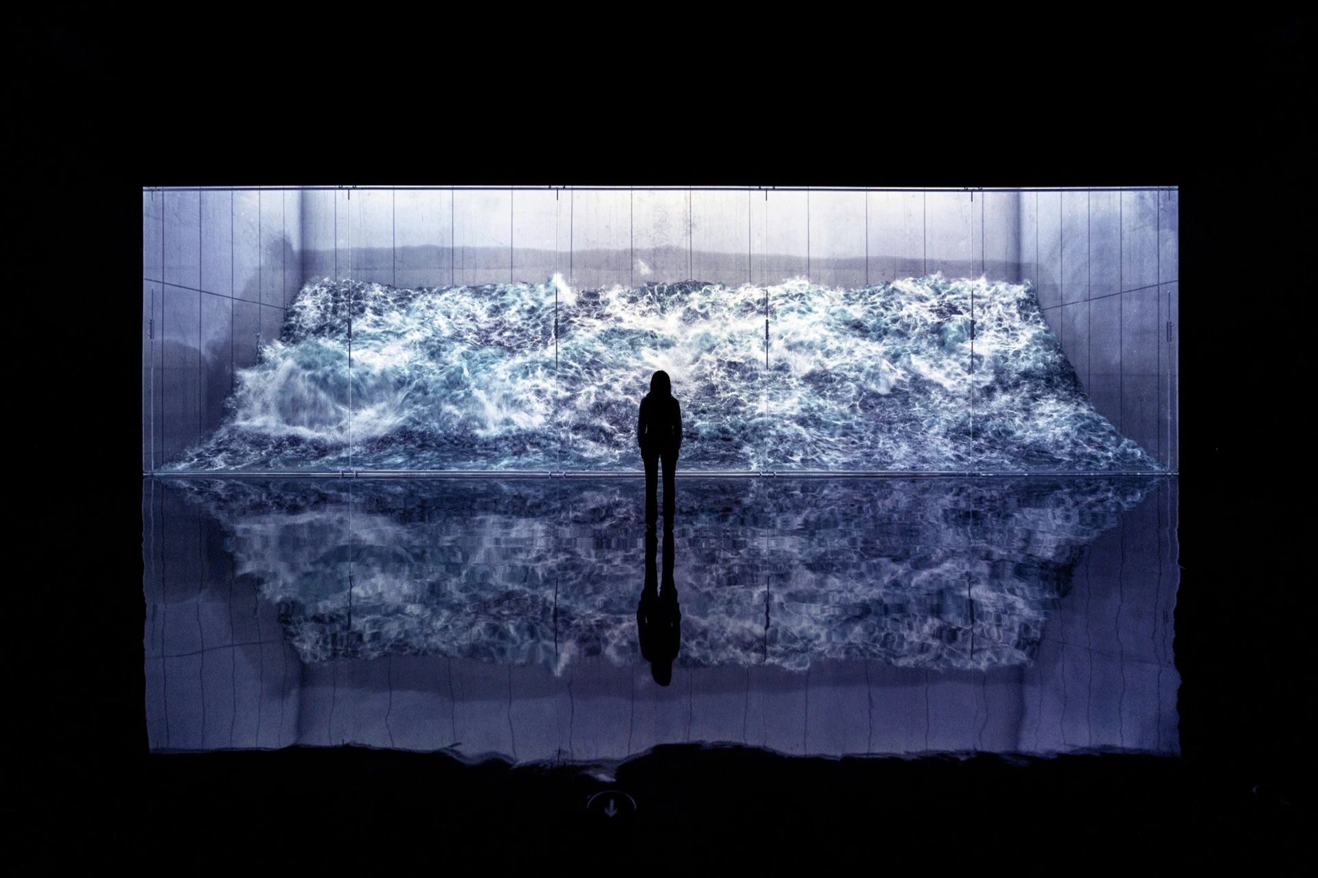 Silhouette of a person standing in front of a large, lit-up digital display of turbulent ocean waves with a reflective floor.