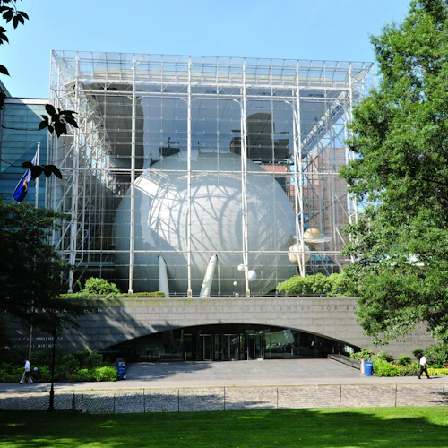 Modernes Gebäude mit einer großen Glasstruktur, in der eine Kugel untergebracht ist, umgeben von Bäumen. Menschen gehen auf dem Weg darunter.