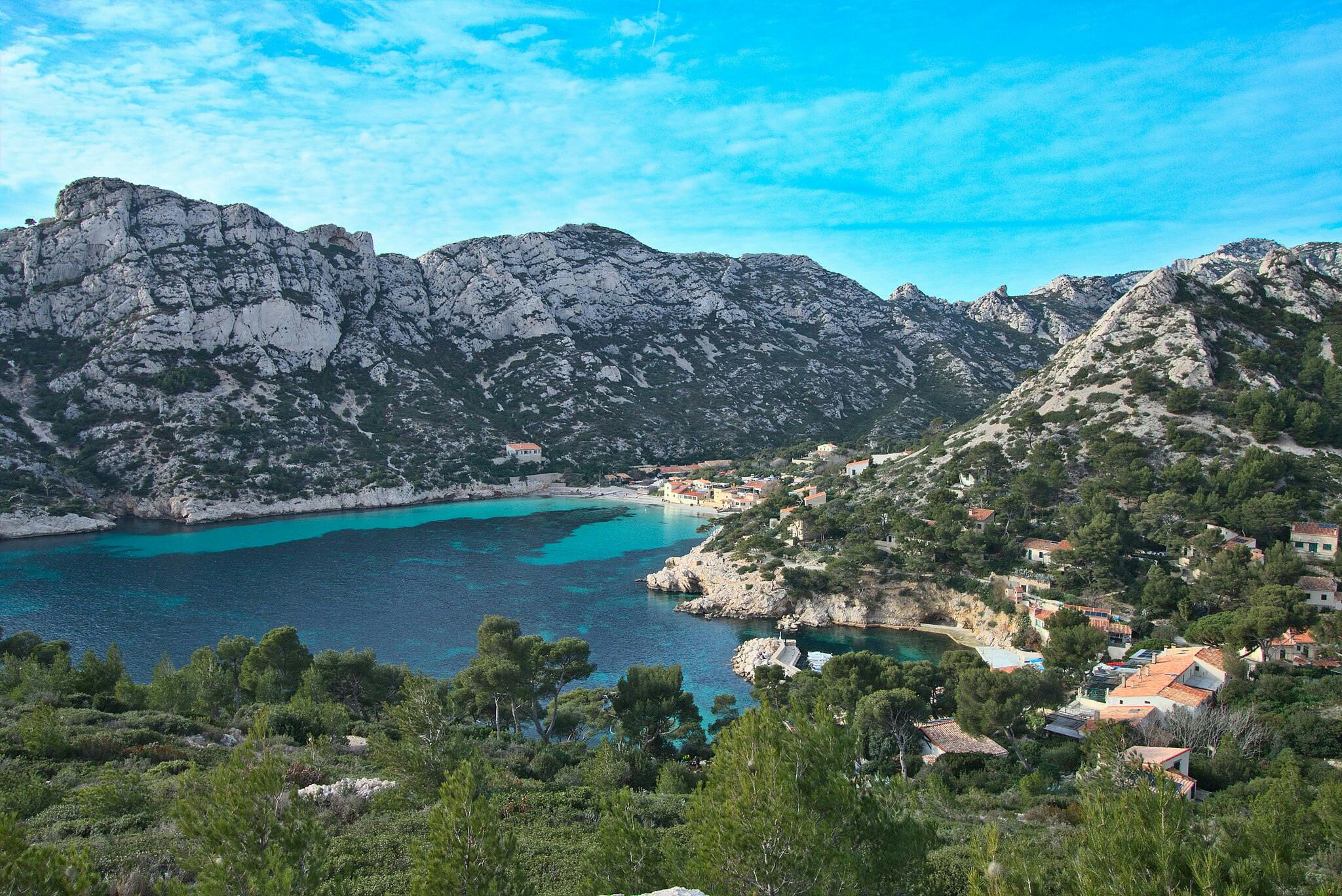 A coastal village nestled between rocky mountains, featuring turquoise waters, scattered houses, and lush green trees under a blue sky.