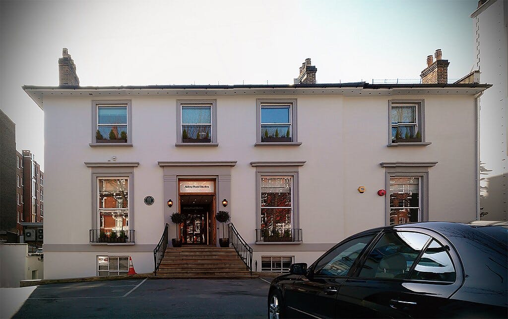 White two-story building with "Abbey Road Studios" sign above the door, four windows, steps leading up, and a black car in front.
