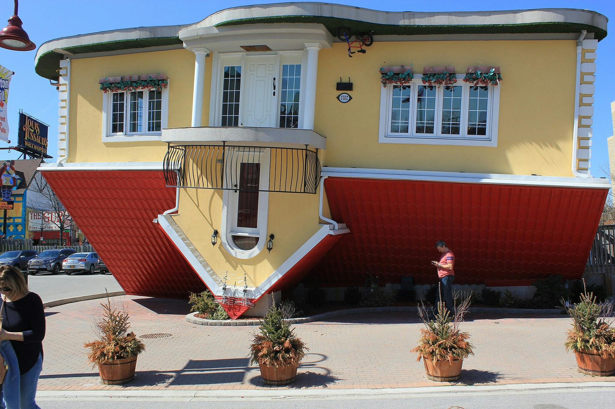 An upside-down house with a red roof and yellow walls on a sunny day, people walking nearby, and a Waxworks sign in the background.