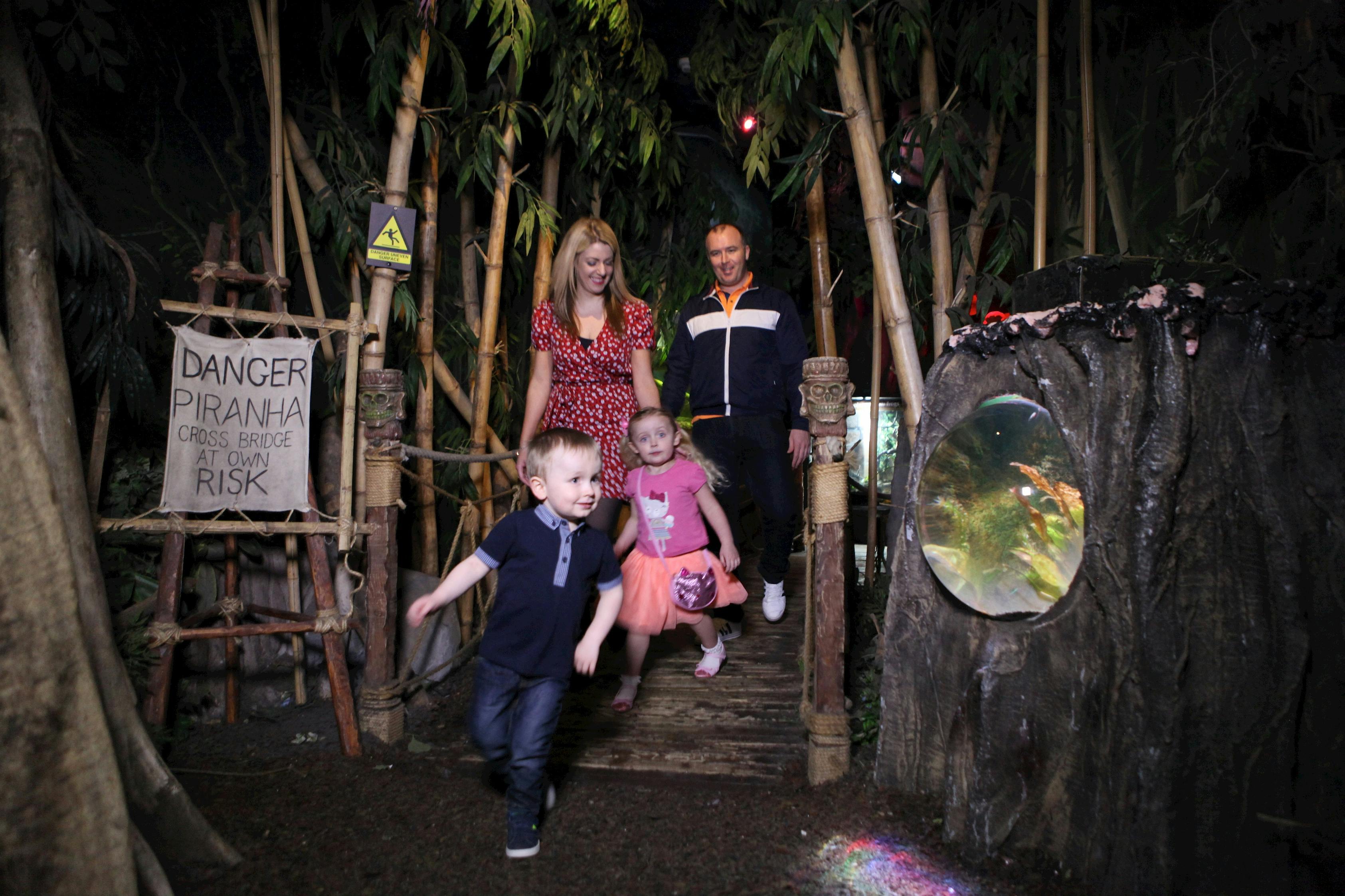 Two children running ahead of two adults on a wooden bridge in a jungle-themed setting with a sign warning about piranhas.