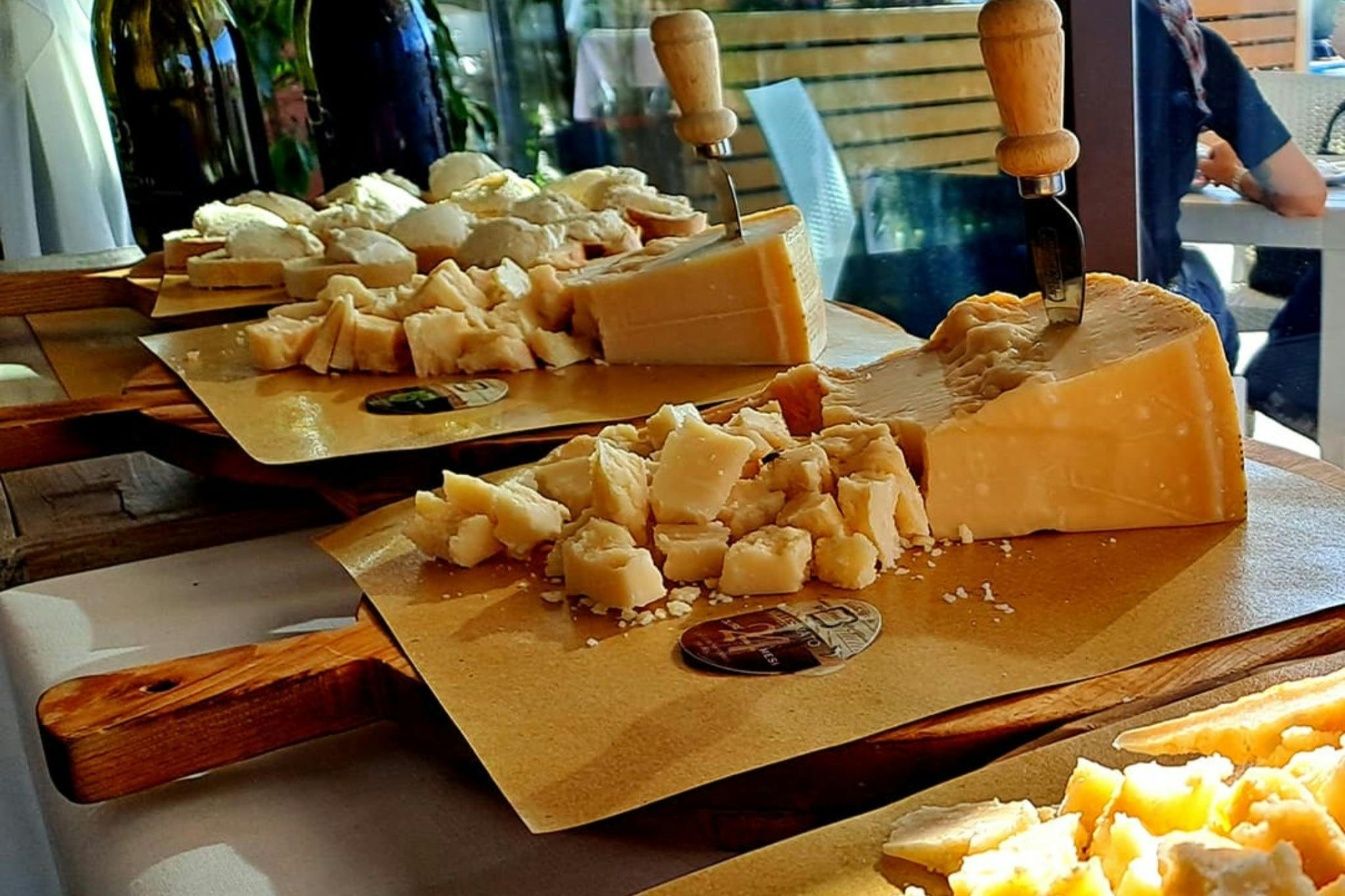 Pezzi di formaggio su un tagliere con coltelli incastrati, disposti su un tavolo. Sullo sfondo una sedia e del verde.