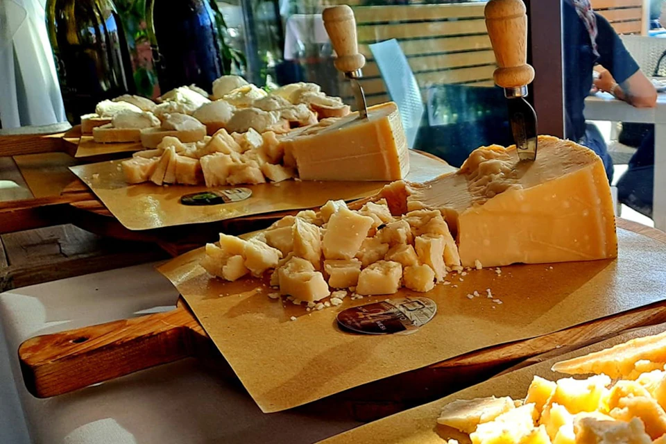 Chunks of cheese on a cutting board with knives embedded, set on a table. Background includes a chair and some greenery.