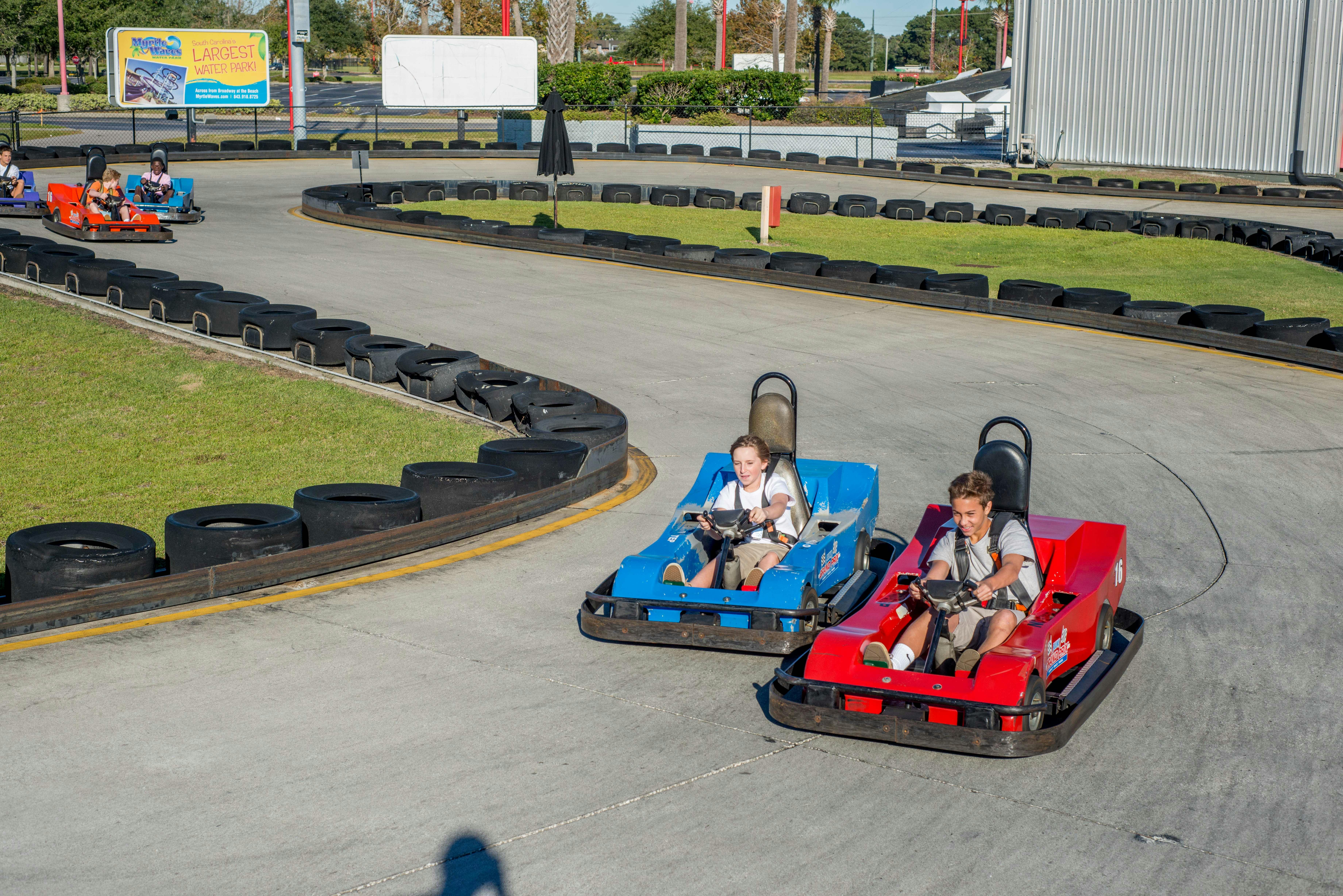 Two people driving go-karts on a track; one in a blue go-kart and the other in a red go-kart, surrounded by tire barriers.