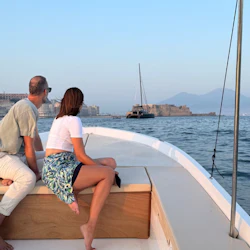 Two people seated on a boat's deck, looking towards a distant cityscape and mountains over calm water.