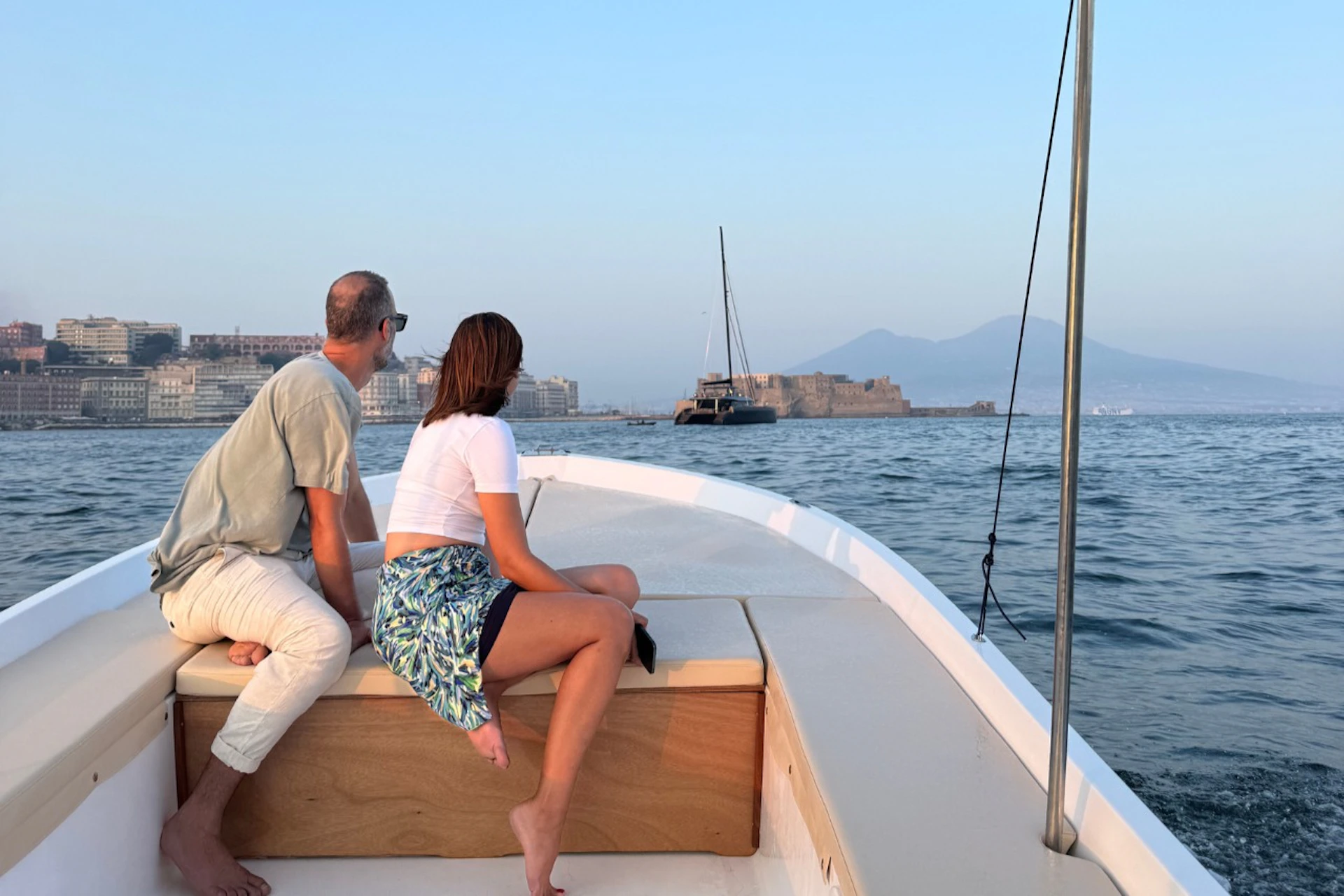 Two people seated on a boat's deck, looking towards a distant cityscape and mountains over calm water.