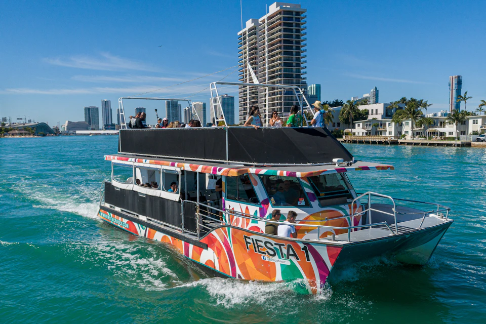 A colorful boat named 'Fiesta 1' on water with passengers aboard, against a backdrop of modern buildings and a blue sky.
