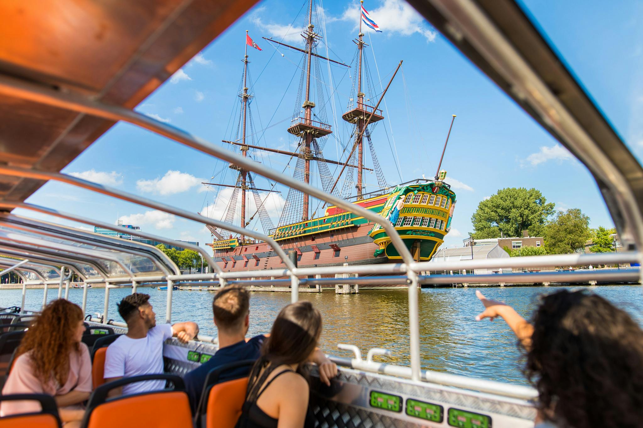 Mensen op een boottocht zien een groot, kleurrijk, historisch zeilschip aangemeerd aan het water onder een strakblauwe hemel.