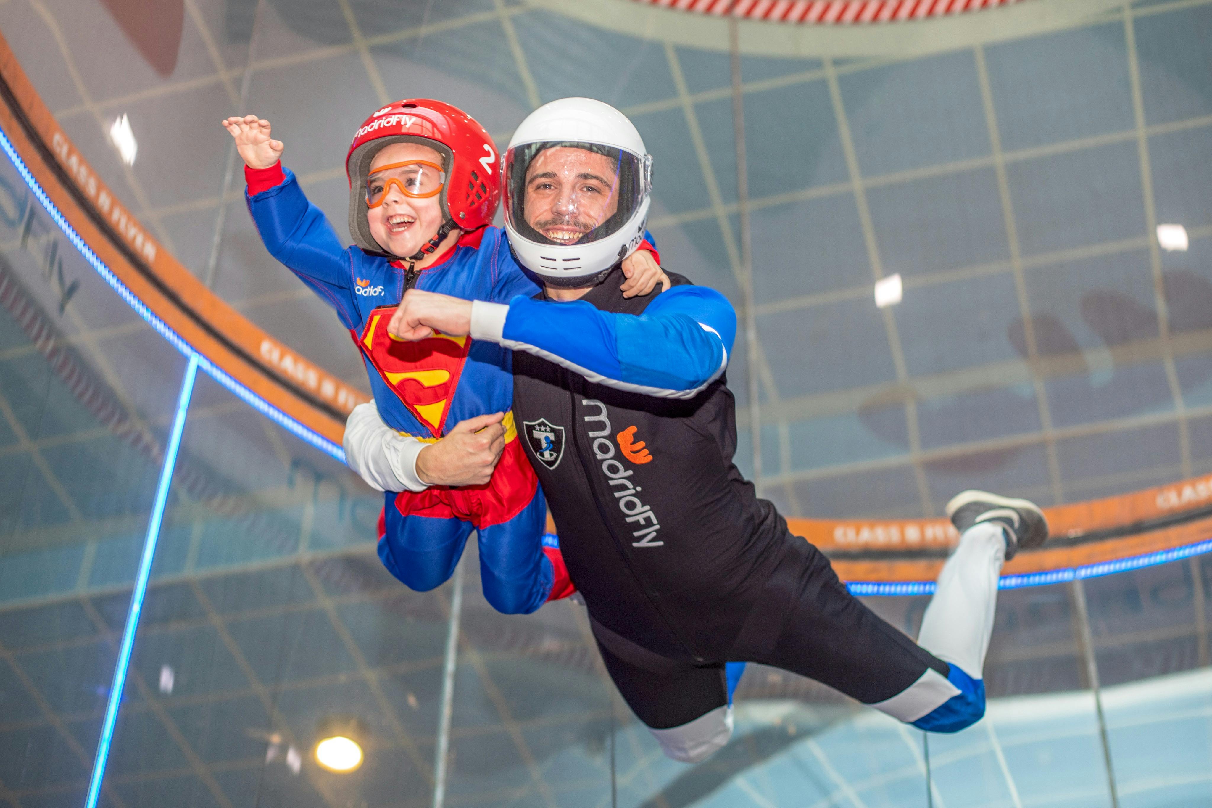 A child in a Superman costume and a man in a flight suit are joyfully indoor skydiving in a wind tunnel.