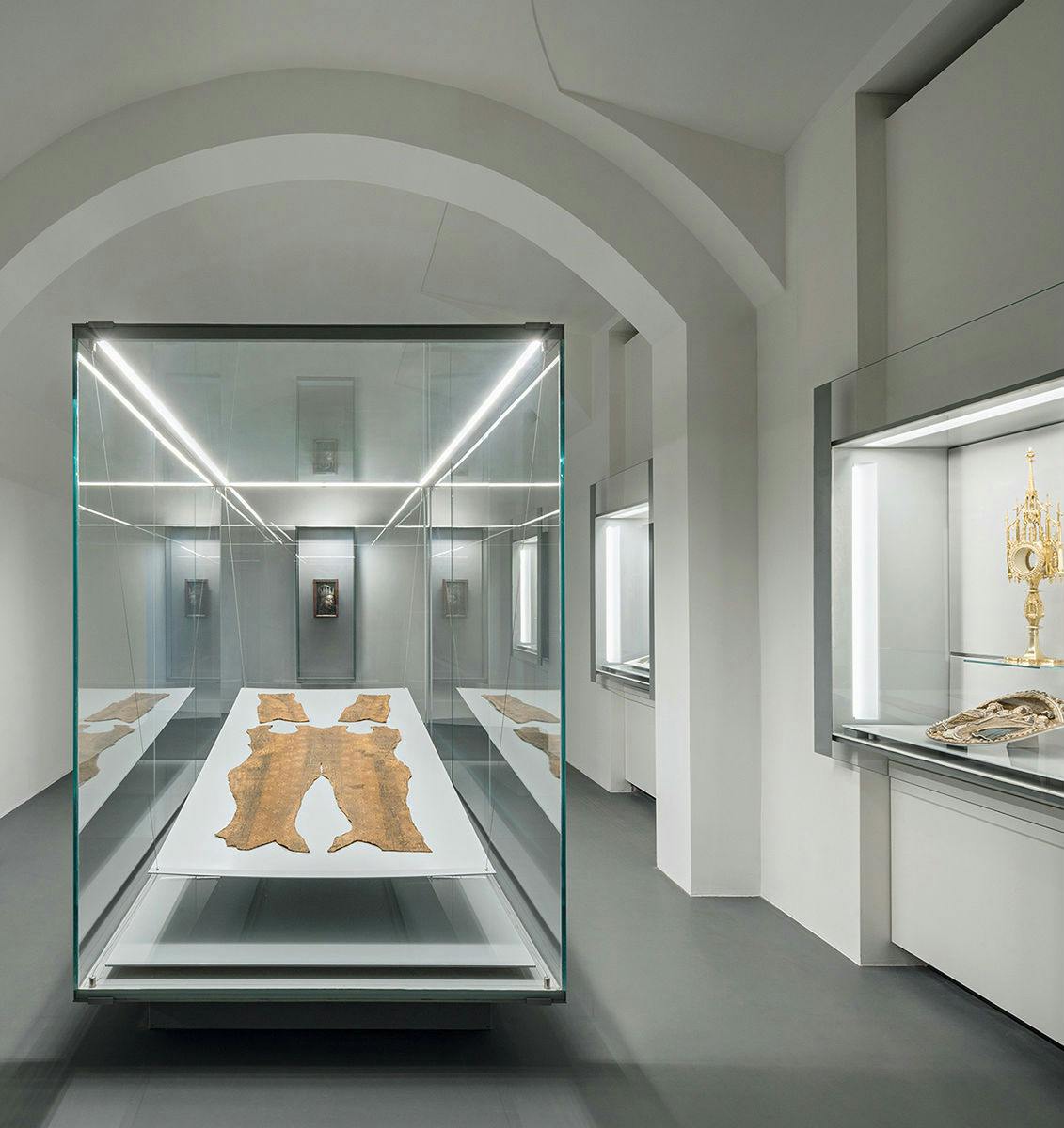 A modern museum room featuring a glass display case with leather artifacts, and other exhibits in illuminated wall cases.