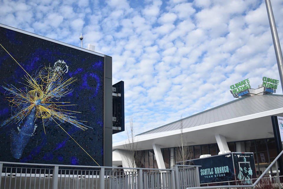 Mosaic artwork on a building, with the Seattle Kraken Team Store in the foreground and a partly cloudy sky above.